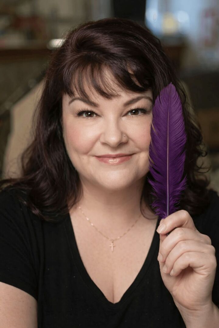 Woman with dark hair holding a purple feather close to her face, smiling, wearing a black top and a delicate necklace.
